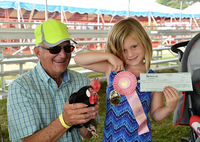 A man holds a rooster and a small girl shows off her ribbon and check for winning the Rooster Crowing Contest at the Carver County Fair