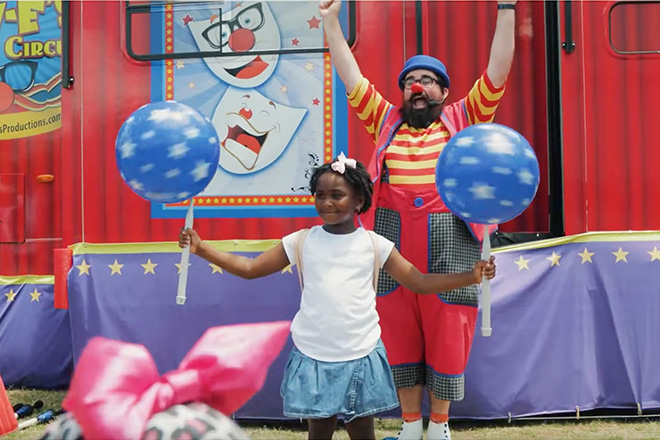 a girl spins two balls on sticks with Lew-E the clown cheering behind her