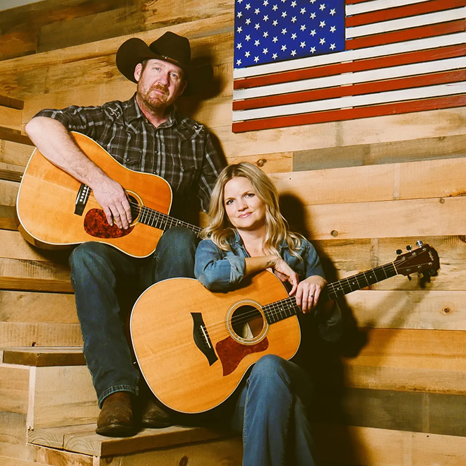 a man and a woman with acoustic guitars pose near a wooden American flag